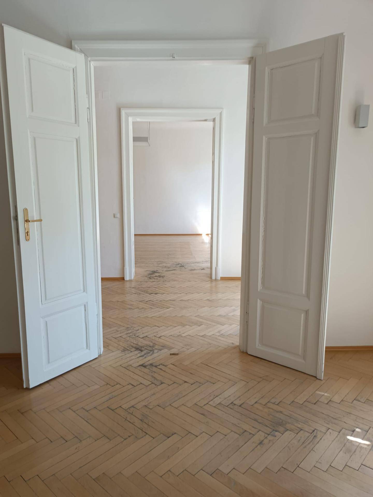 Heller Flur mit Fischgrätparkett und hohen weißen Türen, der in weitere leere Räume führt.
