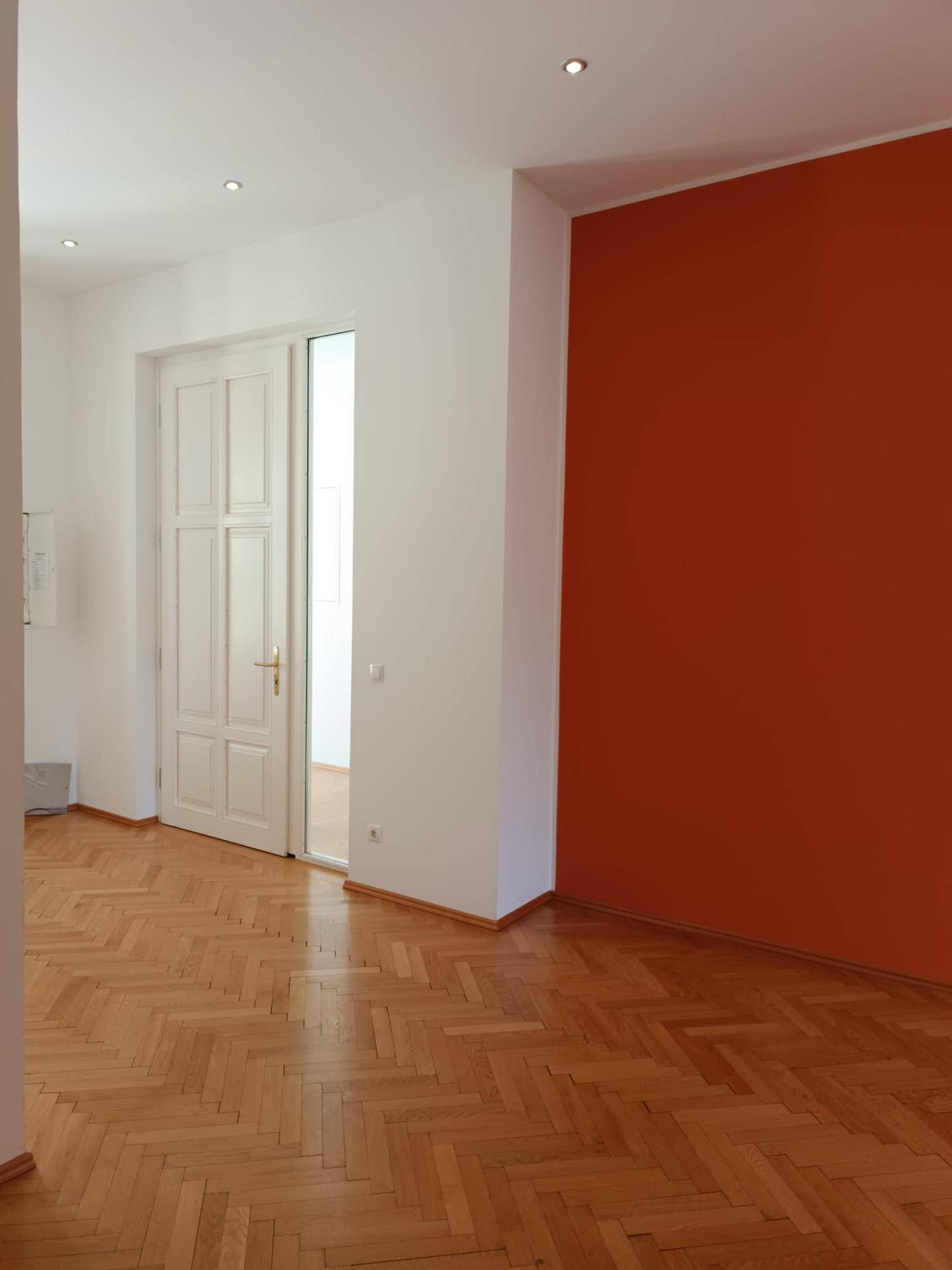Heller Wohnraum mit klassischem Fischgrätparkett und einer auffälligen orangefarbenen Akzentwand.