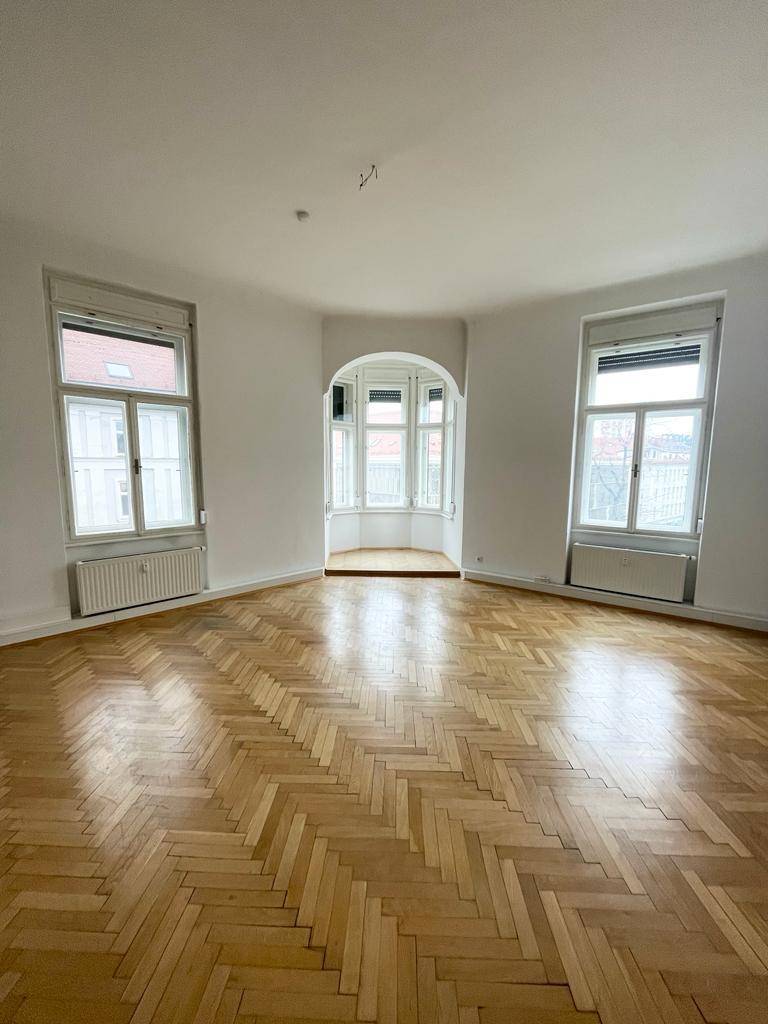 Heller, leerer Wohnraum mit Fischgrätparkett, hohen Decken und einem charmanten Erkerfenster.