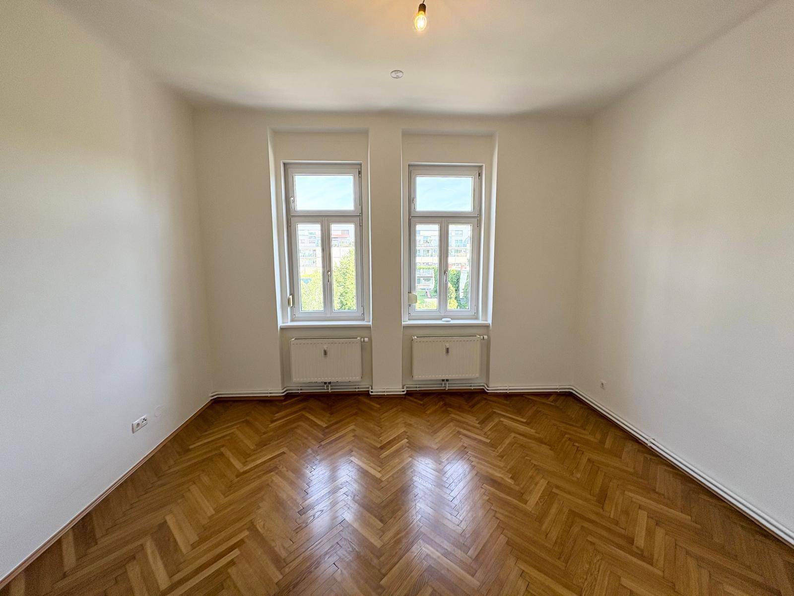 Geräumiges Zimmer mit elegantem Fischgrätparkett und viel Tageslicht durch zwei große Fenster.