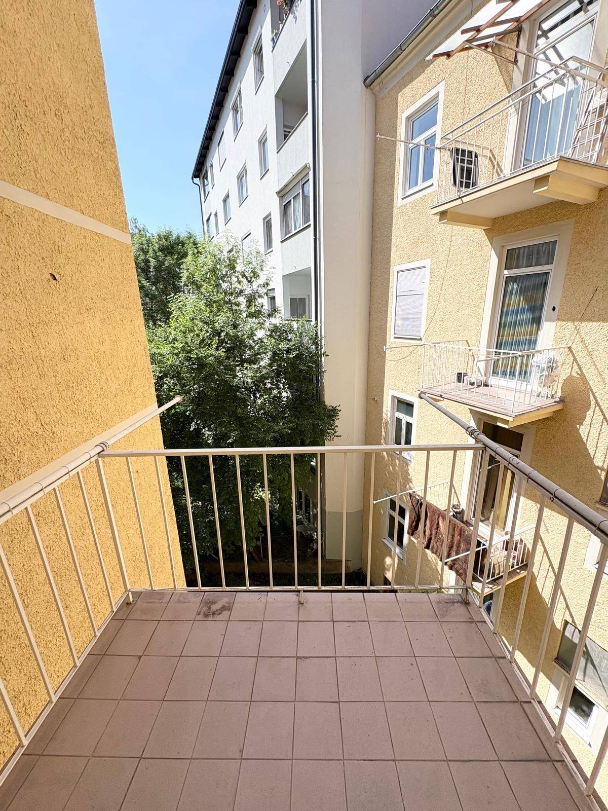 Kleiner Balkon mit Fliesenboden und Blick auf den begrünten Innenhof zwischen den Gebäuden.