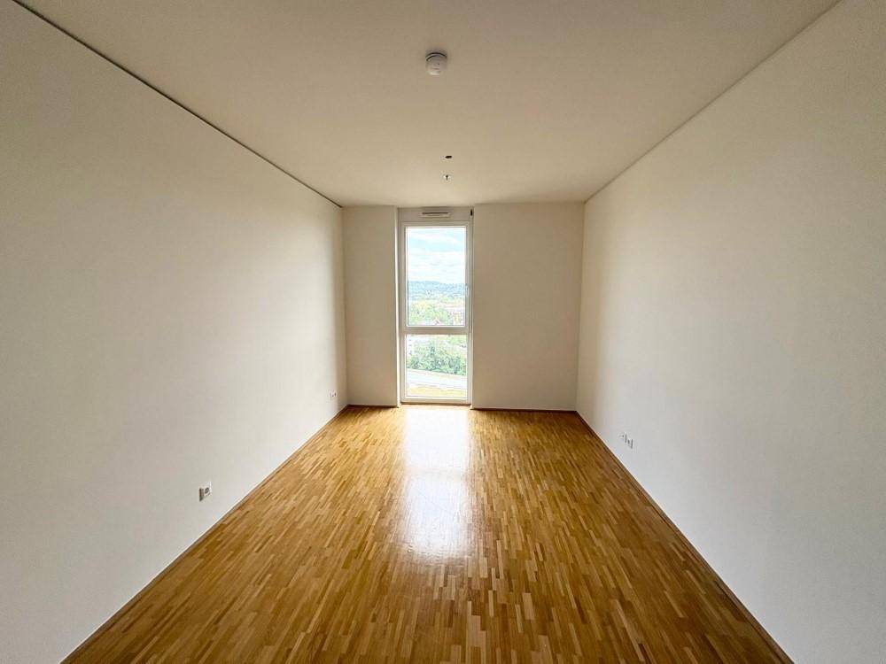 Helles, leeres Zimmer mit Parkettboden und einem hohen Fenster mit Blick ins Grüne.