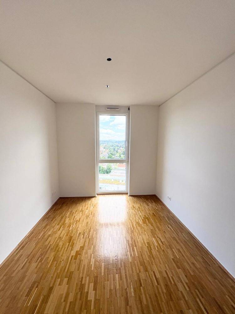 Lichtdurchflutetes Zimmer mit Holzboden und einem hohen Fenster, das eine schöne Aussicht ermöglicht.