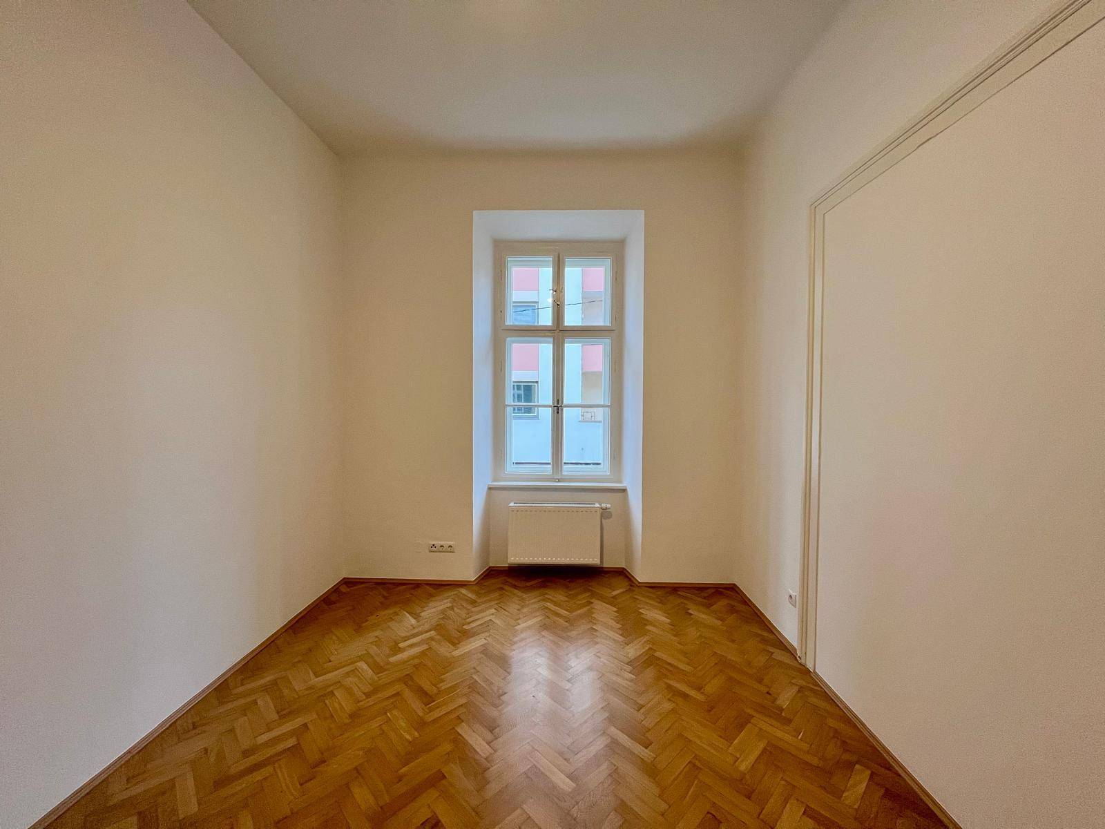 Helles Zimmer mit Fischgrätparkett und einem Fenster, ideal als Schlafzimmer nutzbar.