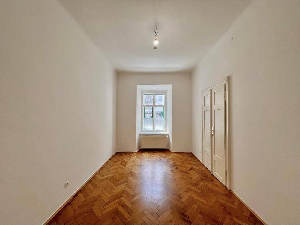 Leeres Zimmer mit Fischgrätparkett, hohen Decken und einem Fenster, bereit zur Einrichtung.