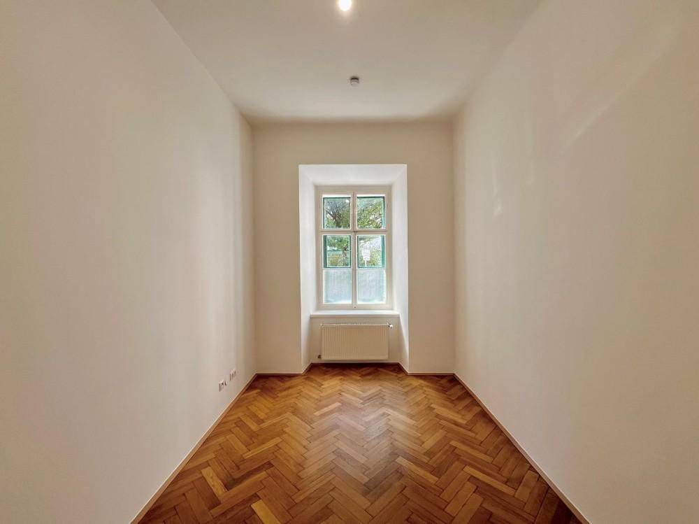 Helles Zimmer mit klassischem Fischgrätparkett und einem Fenster, ideal als Schlaf- oder Arbeitszimmer.