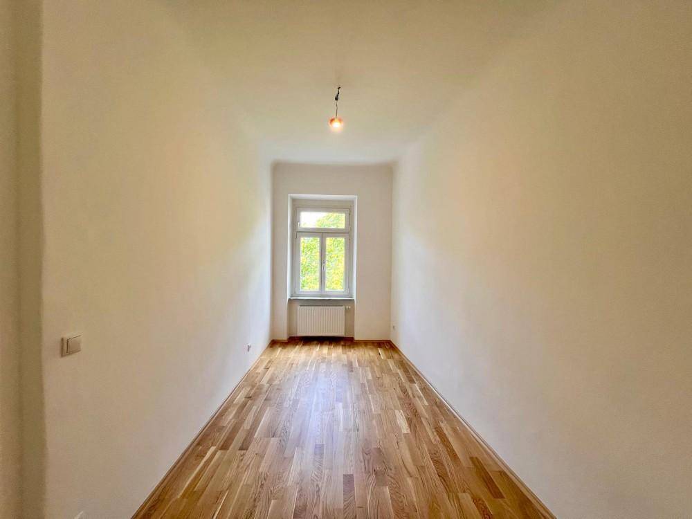 Schmaler Raum mit hellem Parkettboden, weißen Wänden und einem Fenster mit Heizkörper.