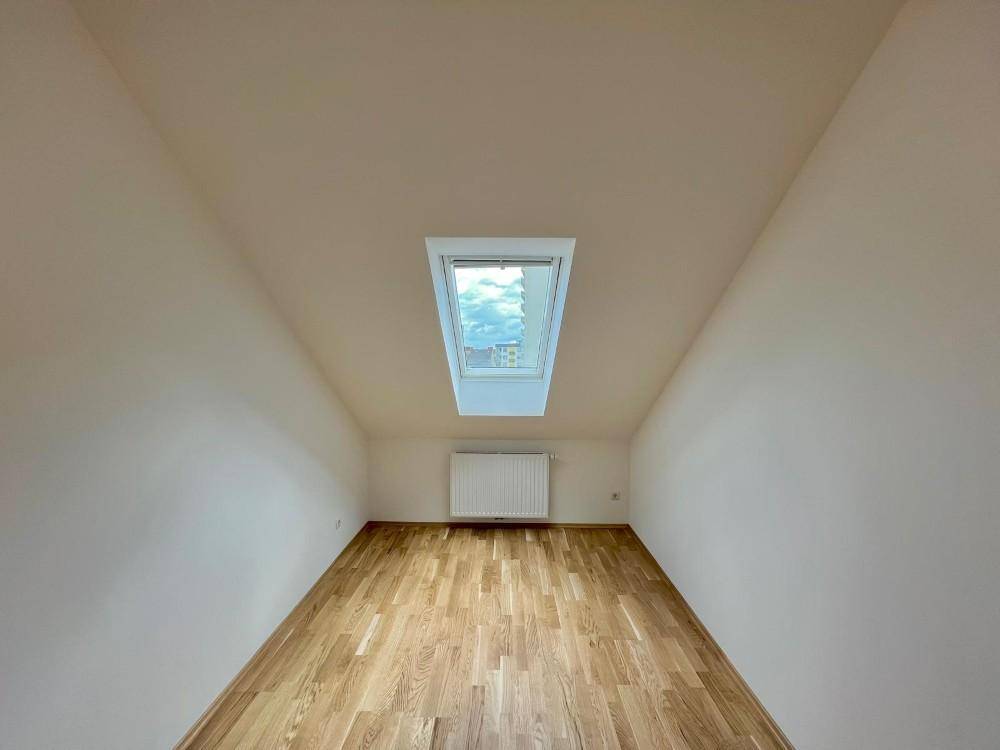 Helles Zimmer im Dachgeschoss mit schrägen Wänden, Parkettboden und einem Dachfenster.
