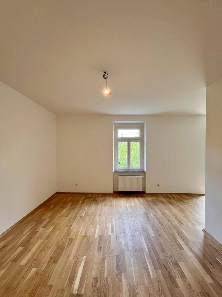Helles, leeres Zimmer mit Parkettboden, weißen Wänden und einem Fenster mit Heizkörper.