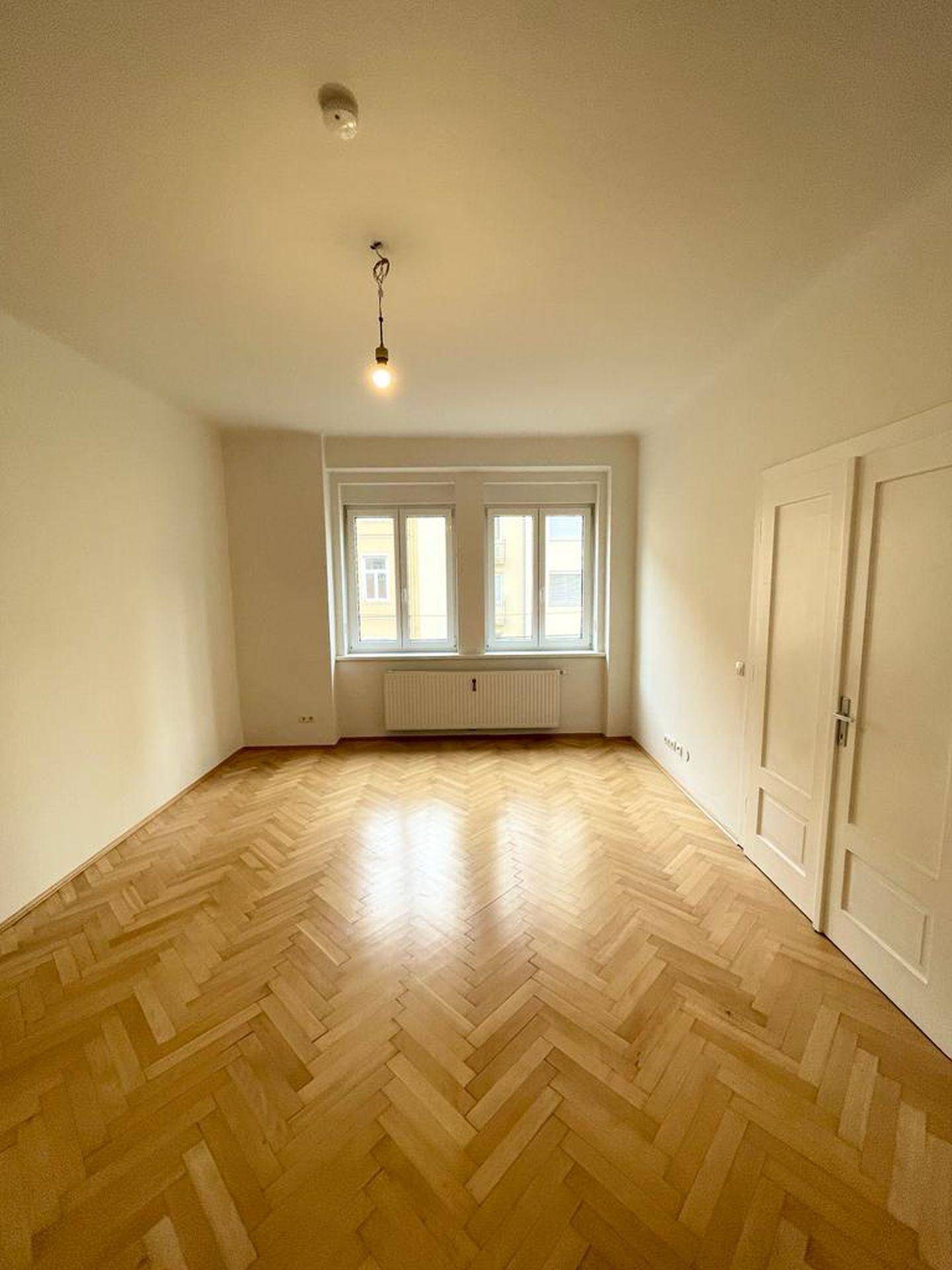 Geräumiges Zimmer mit Fischgrätparkett, zwei Fenstern und weißen Türen.