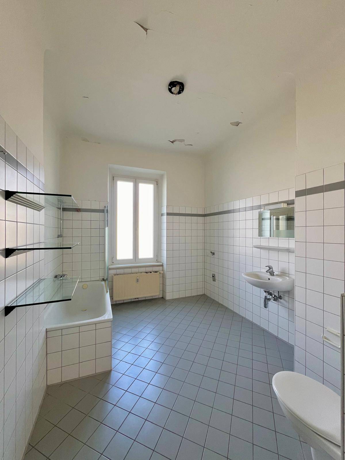 Helles Badezimmer mit Badewanne, Waschbecken und Fenster, gefliest in Weiß und Grau.