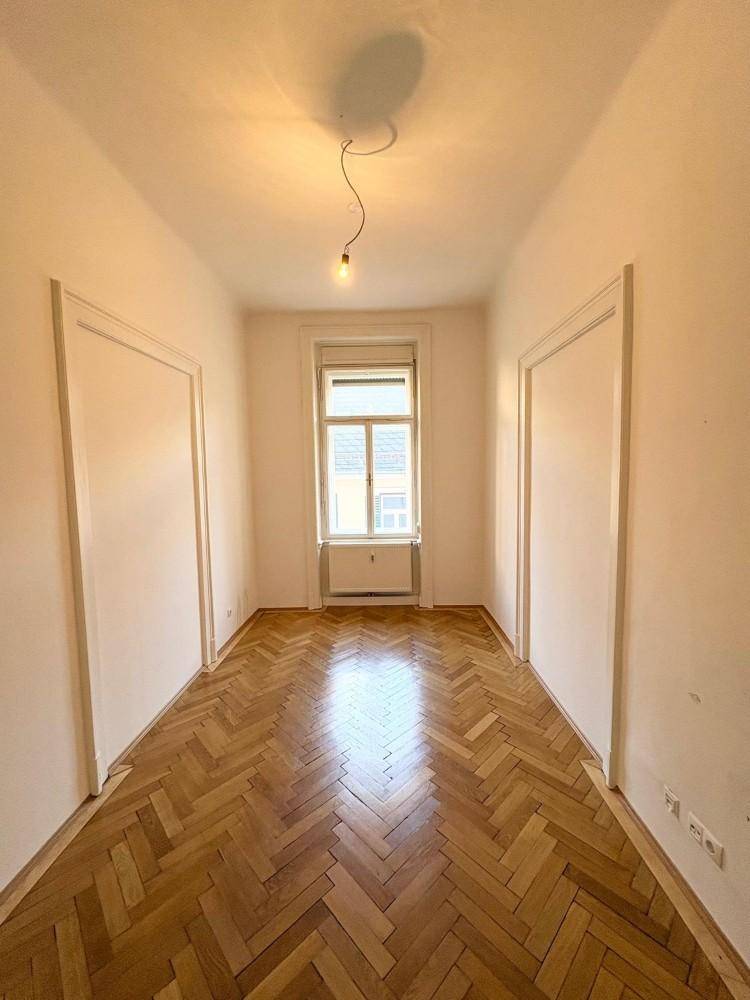Langer, heller Raum mit Fischgrätparkett und einem Fenster, ideal als Flur oder kleines Zimmer.