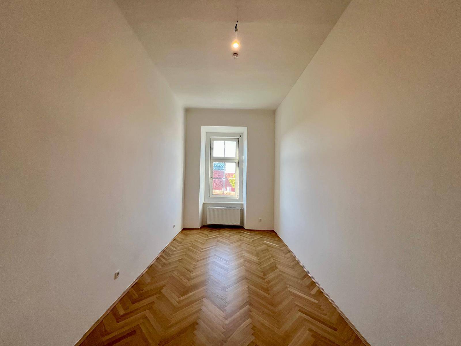 Helles Zimmer mit klassischem Fischgrätparkett und einem Fenster, das viel Licht hereinlässt.