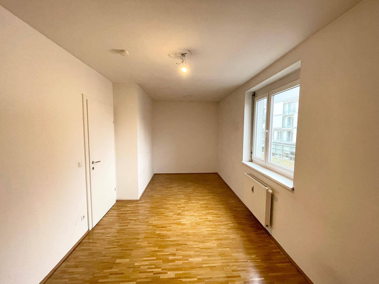 Leeres Schlafzimmer mit Parkettboden, großem Fenster und viel Tageslicht.