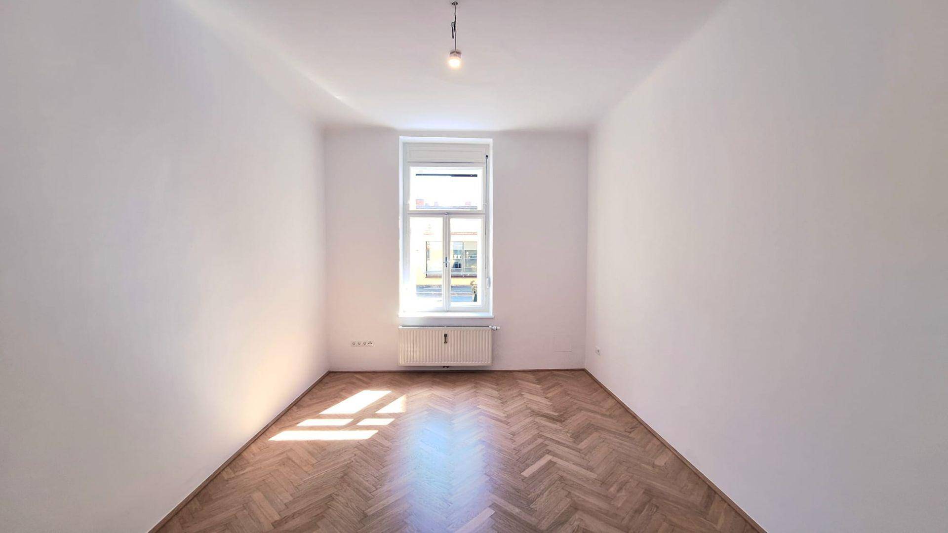 Helles, leeres Zimmer mit Fischgrätparkett und einem Fenster, das den Raum erhellt.