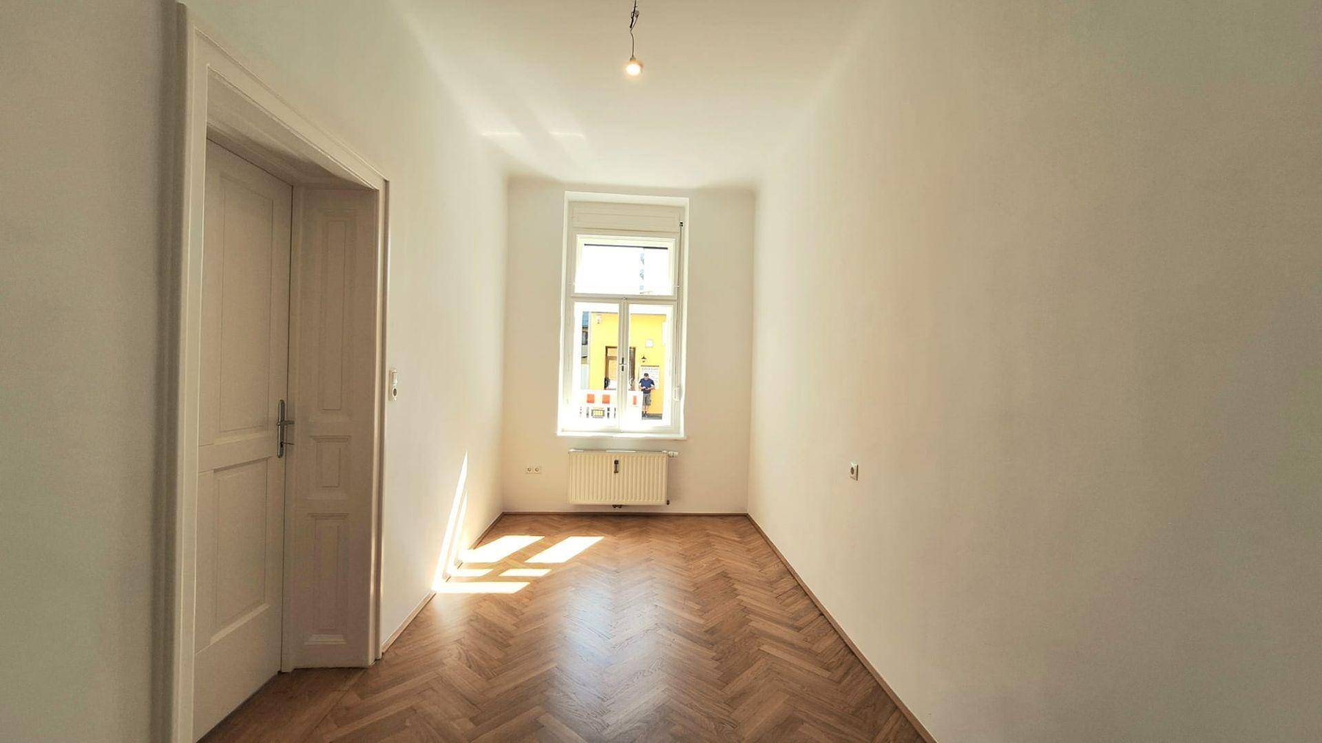 Leeres Zimmer mit klassischem Fischgrätparkett und einem Fenster, das viel Licht hereinlässt.