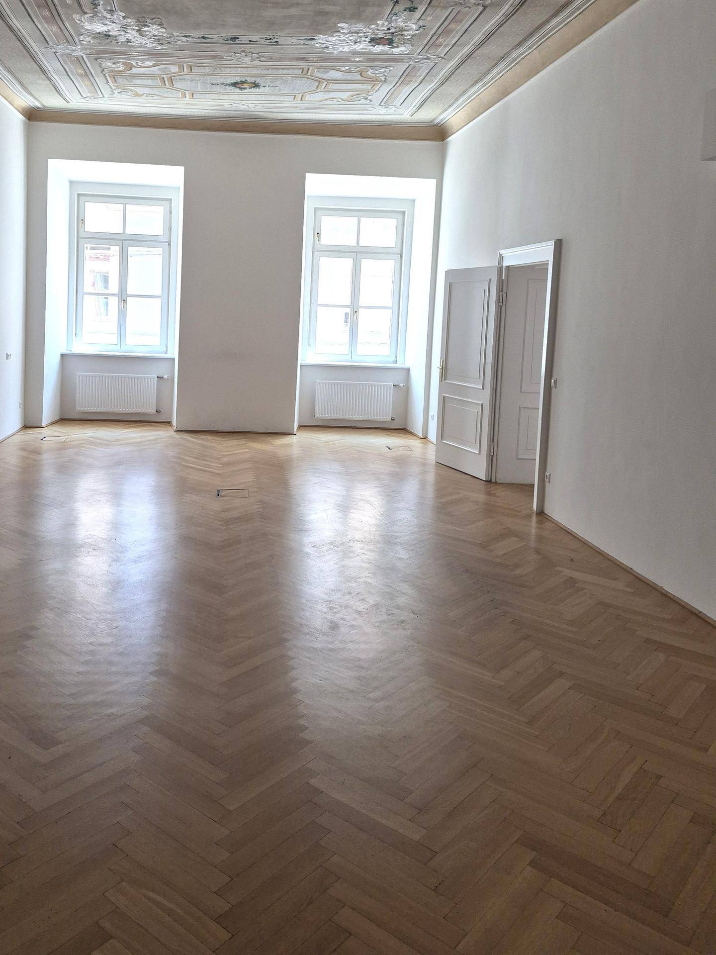 Geräumiger Raum mit Fischgrätparkett, hohen Decken und zwei großen Fenstern, die viel Tageslicht hereinlassen.