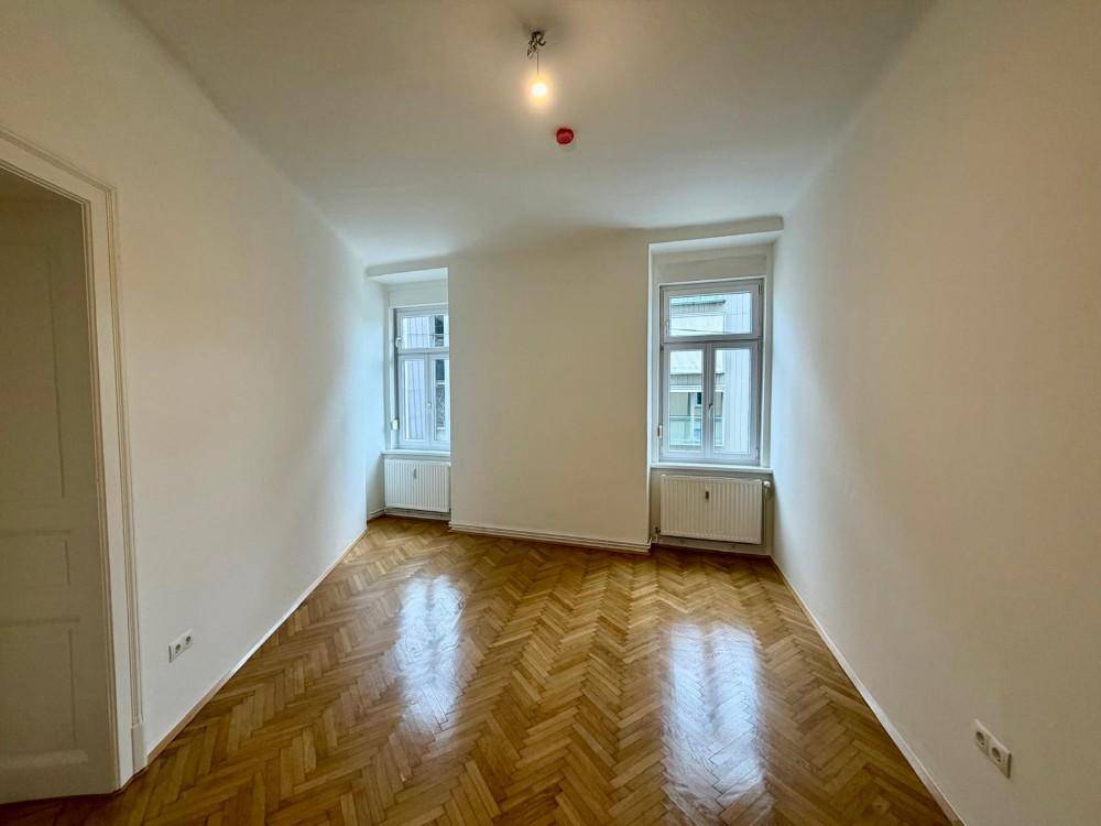 Helles, leeres Zimmer mit Fischgrätparkettboden und zwei Fenstern, gut als Schlafzimmer geeignet.