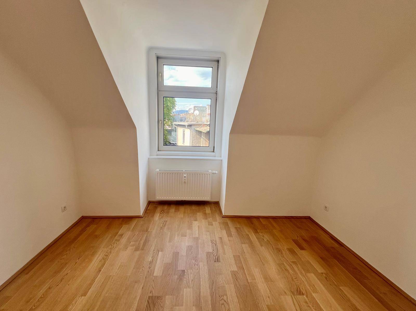 Helles Dachzimmer mit Holzboden und einem Fenster, das viel Tageslicht hereinlässt.