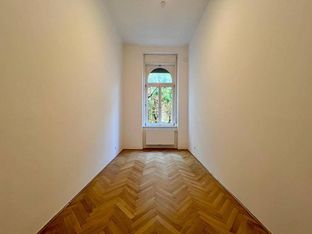 Geräumiges Zimmer mit hellem Fischgrätparkett und einem großen Fenster, das viel Tageslicht hereinlässt.