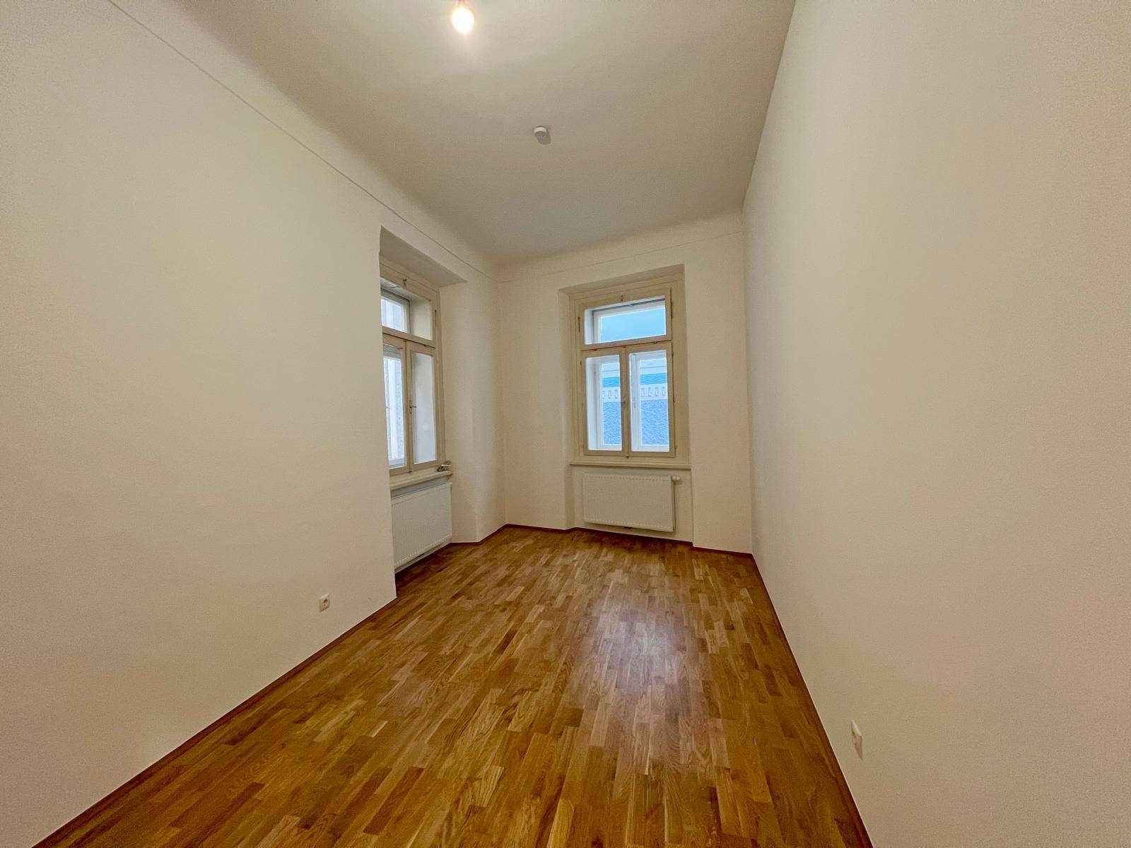 Geräumiges Zimmer mit hellem Parkettboden, weißen Wänden und zwei Fenstern für viel Tageslicht.