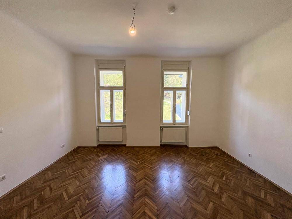 Geräumiges Zimmer mit elegantem Fischgrätparkett und hohen Fenstern, die eine helle und einladende Atmosphäre schaffen.