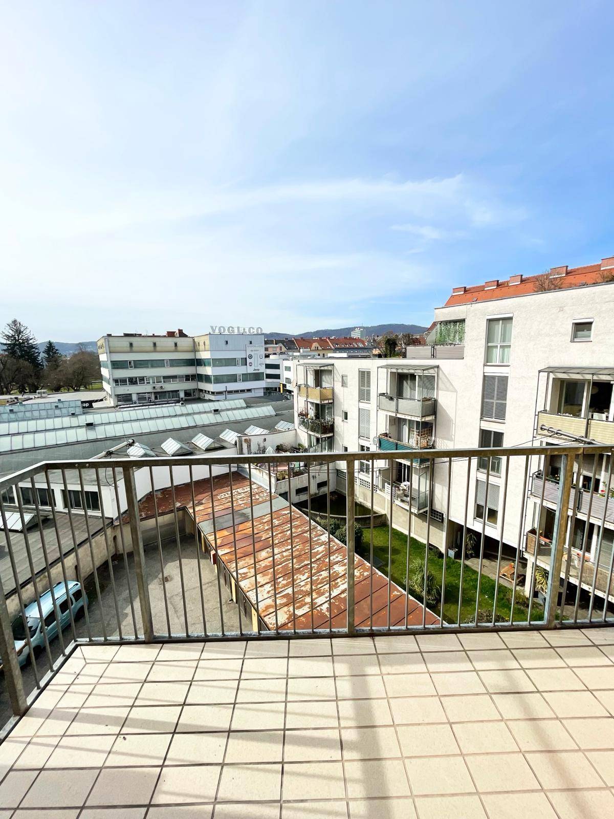 Balkon mit Fliesenboden und Metallgeländer bietet Blick auf umliegende Gebäude und Grünflächen.