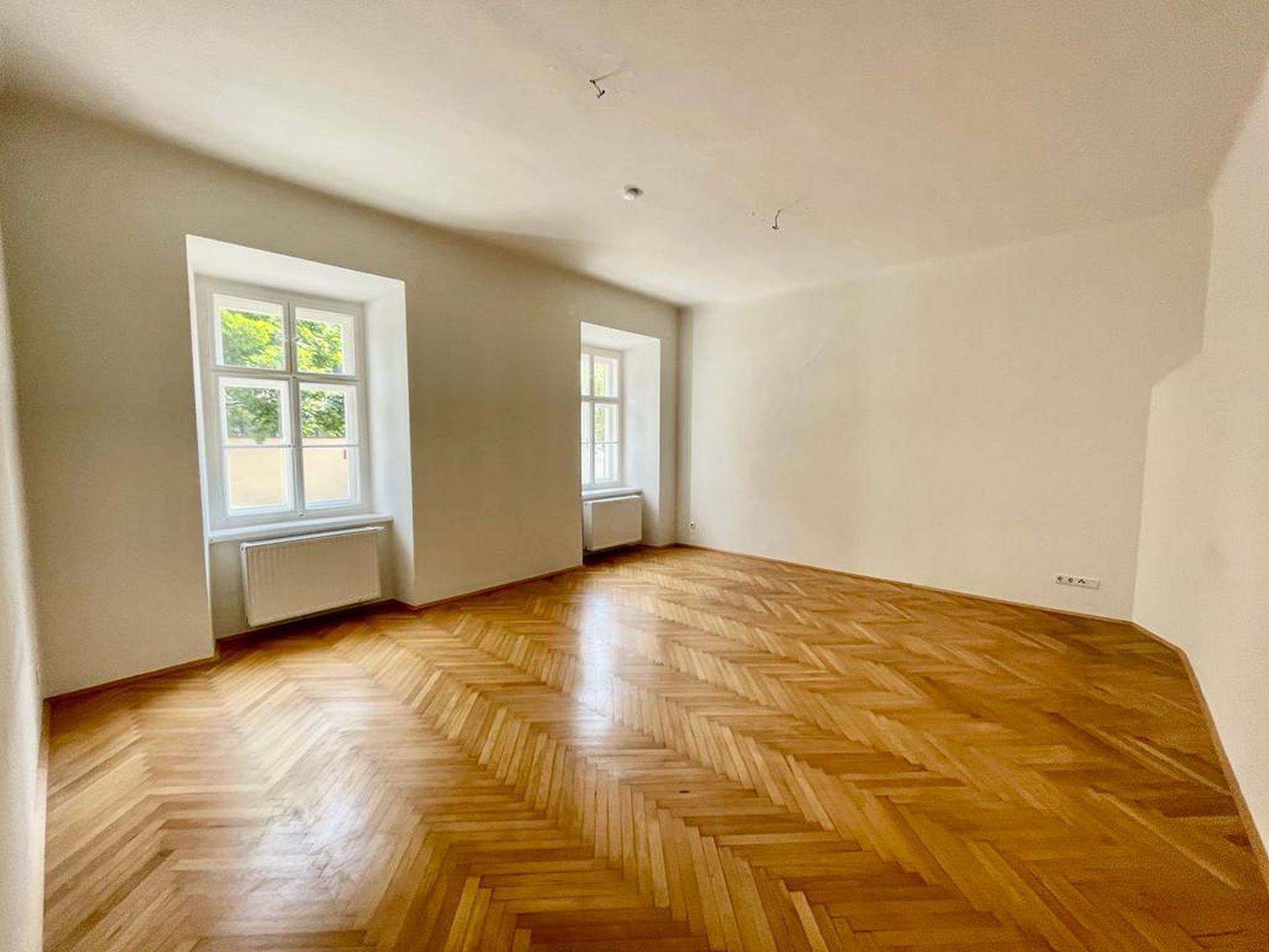 Geräumiges Wohnzimmer mit Fischgrätparkett, hohen Decken und zwei großen Fenstern.