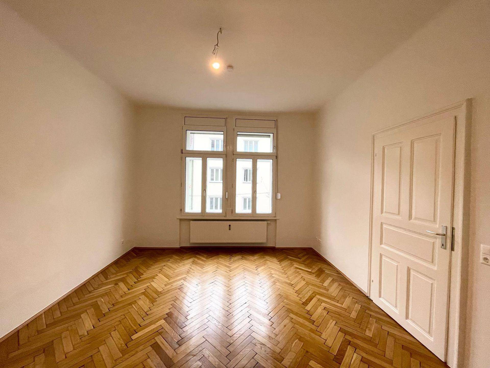 Geräumiges Zimmer mit klassischem Fischgrätparkett und viel Tageslicht durch große Fenster.