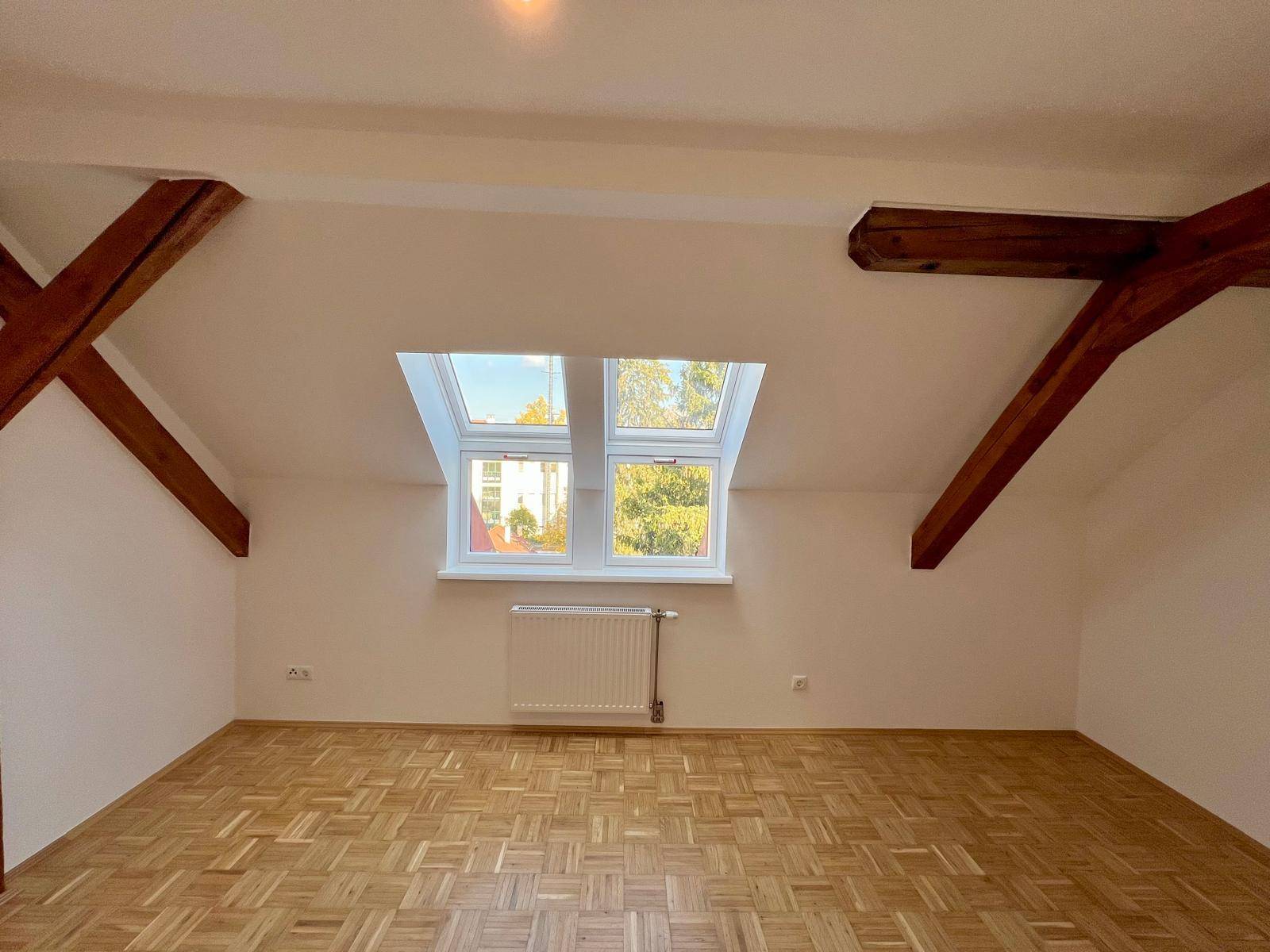 Heller Dachgeschossraum mit zwei Dachfenstern, Parkettboden und Holzbalken.