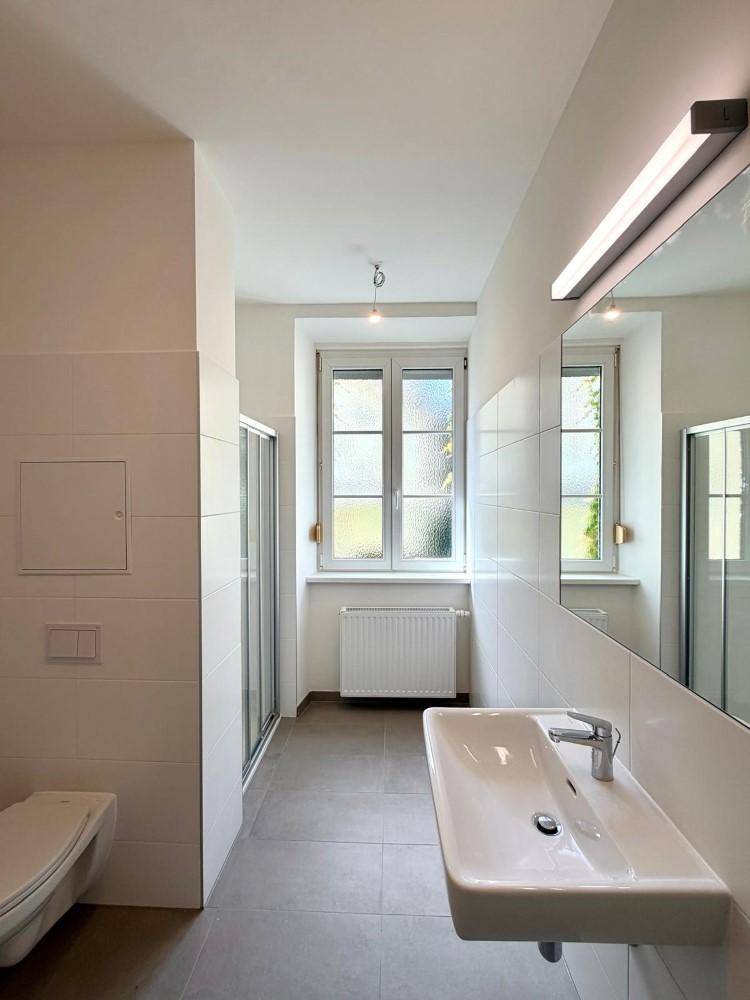 Modernes Badezimmer mit weißen Fliesen, Dusche, Waschbecken und Fenster für Tageslicht.