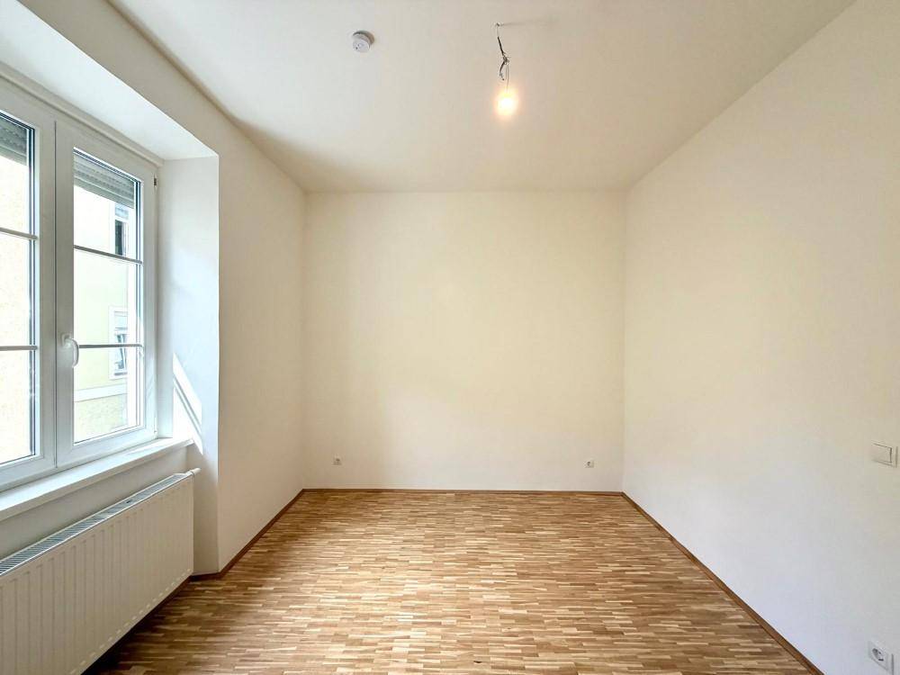 Leerer, heller Raum mit Holzfußboden und großem Fenster, bereit zur individuellen Gestaltung.