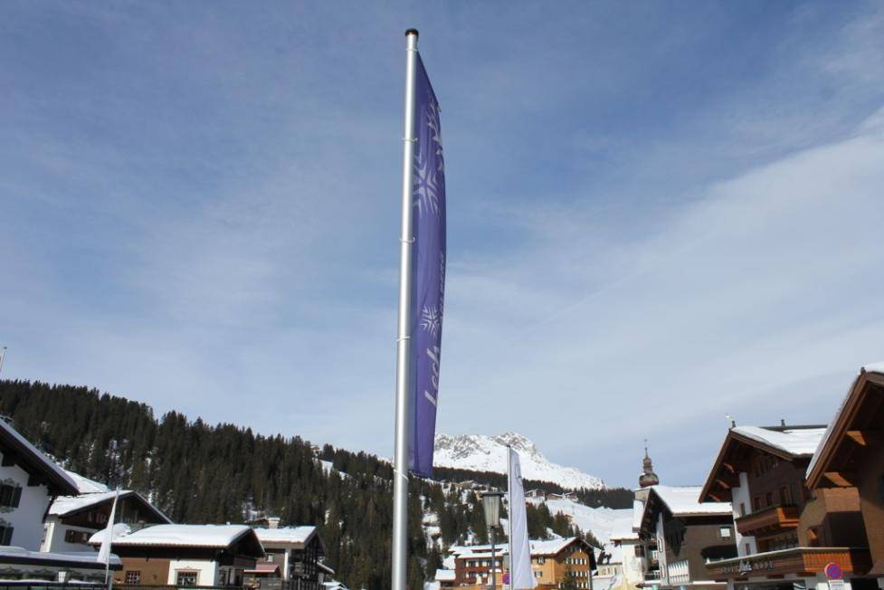 Lila Werbeflagge weht in einem verschneiten Bergdorf unter blauem Himmel mit schneebedeckten Gipfeln.
