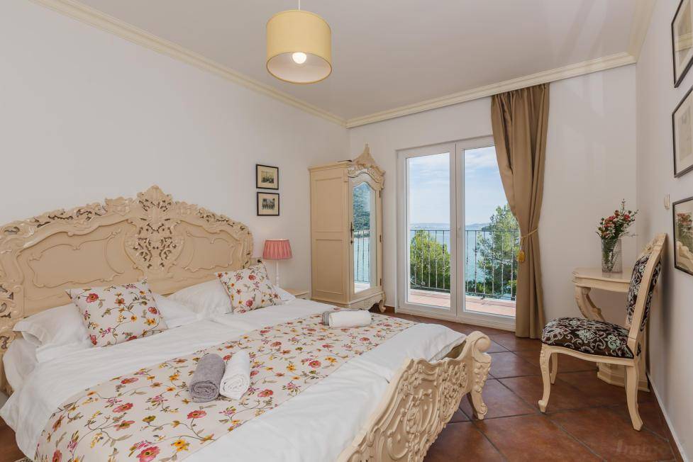 Helles Schlafzimmer mit kunstvollem Doppelbett, floraler Bettwäsche und Balkonzugang mit Meerblick.