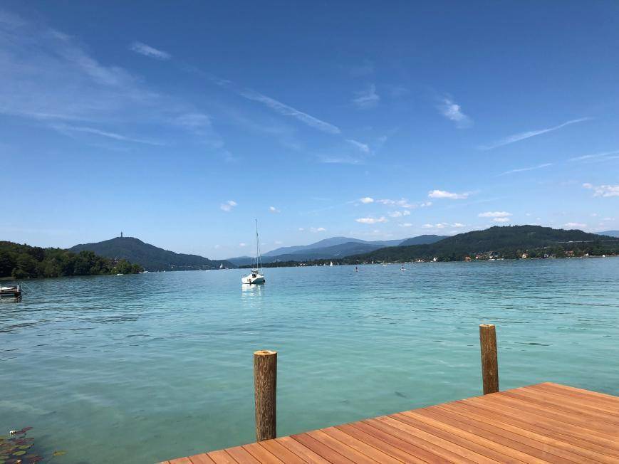 Weitläufiger Blick über den See mit Segelbooten und bewaldeten Bergen unter blauem Himmel.