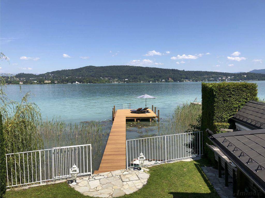 Panoramablick vom Garten auf den See mit privatem Holzsteg, Liegestühlen und Sonnenschirm.