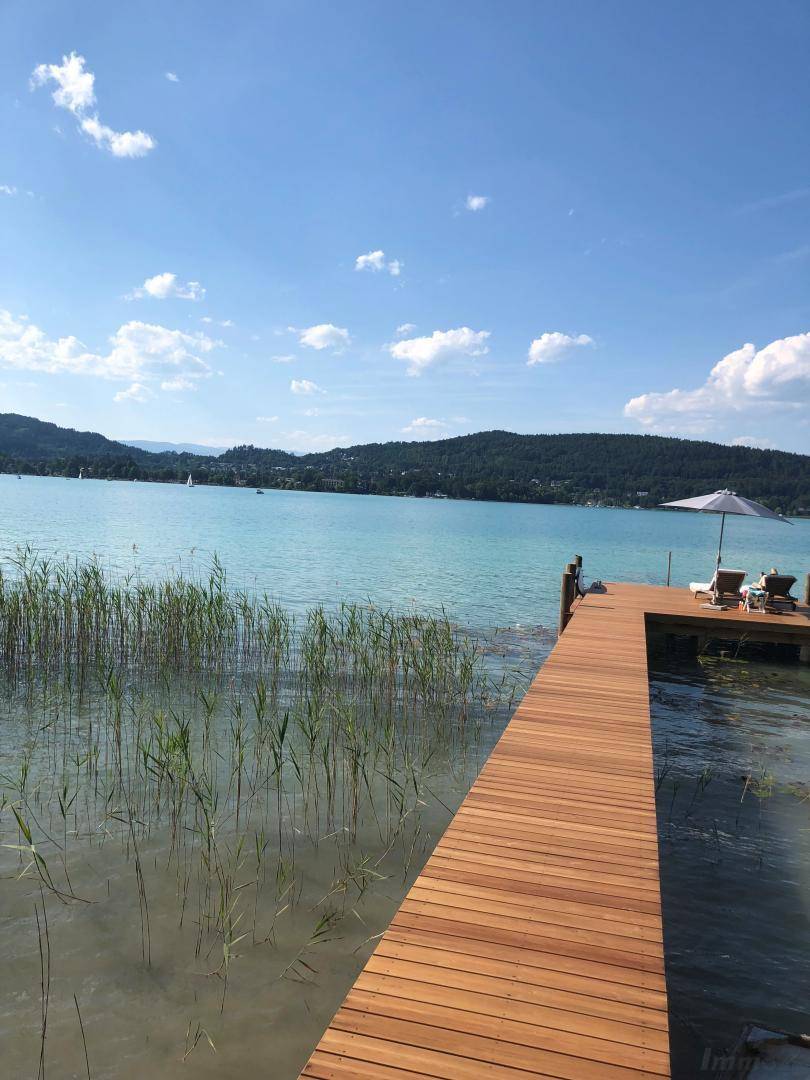 Langer Holzsteg führt in den See, umgeben von Schilf und mit Liegestühlen am Ende.