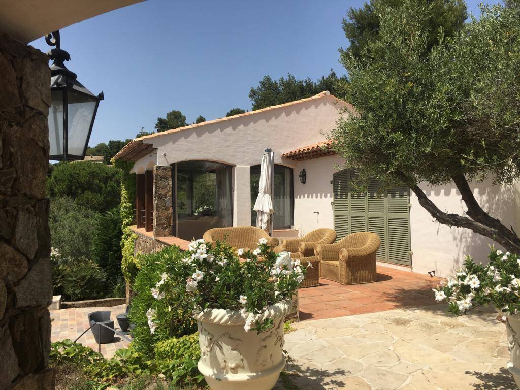 Mediterranes Haus mit heller Fassade und einer möblierten Terrasse, umgeben von üppiger Vegetation.