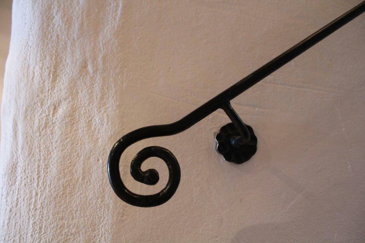Detailaufnahme eines kunstvoll geschmiedeten schwarzen Handlaufs an einer strukturierten weißen Wand.