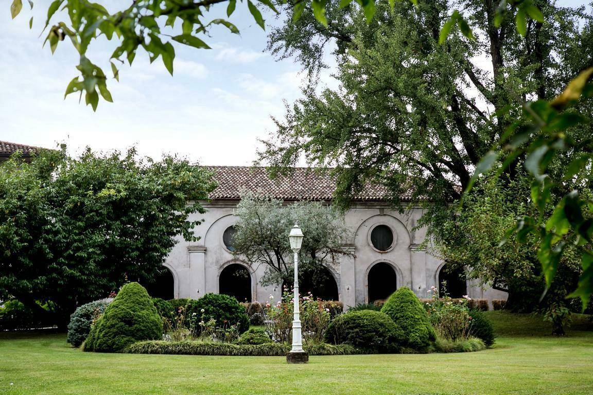 Historisches Gebäude mit Rundbögen und ovalen Fenstern, umgeben von einem gepflegten Garten mit Ziersträuchern.