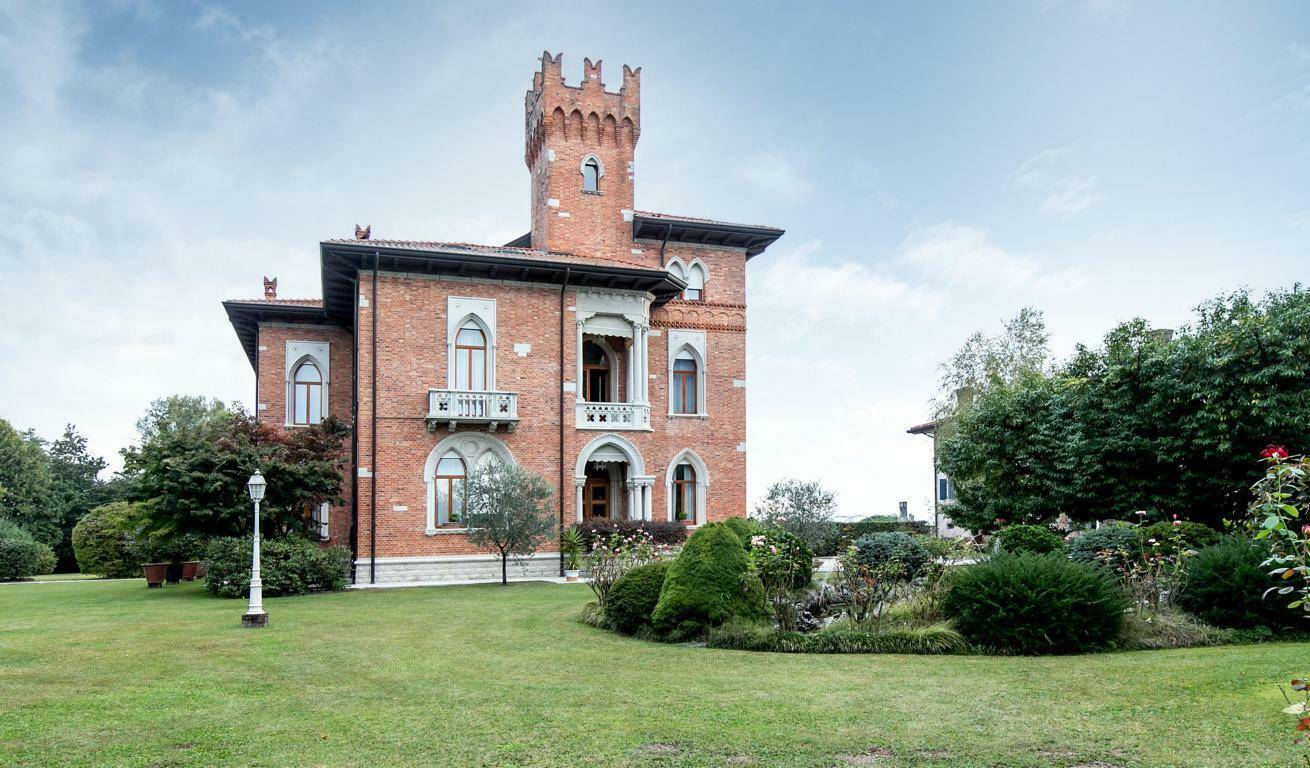 Weitläufiger Rasenbereich vor der historischen Villa mit Ziegelfassade und Turm, umgeben von altem Baumbestand.