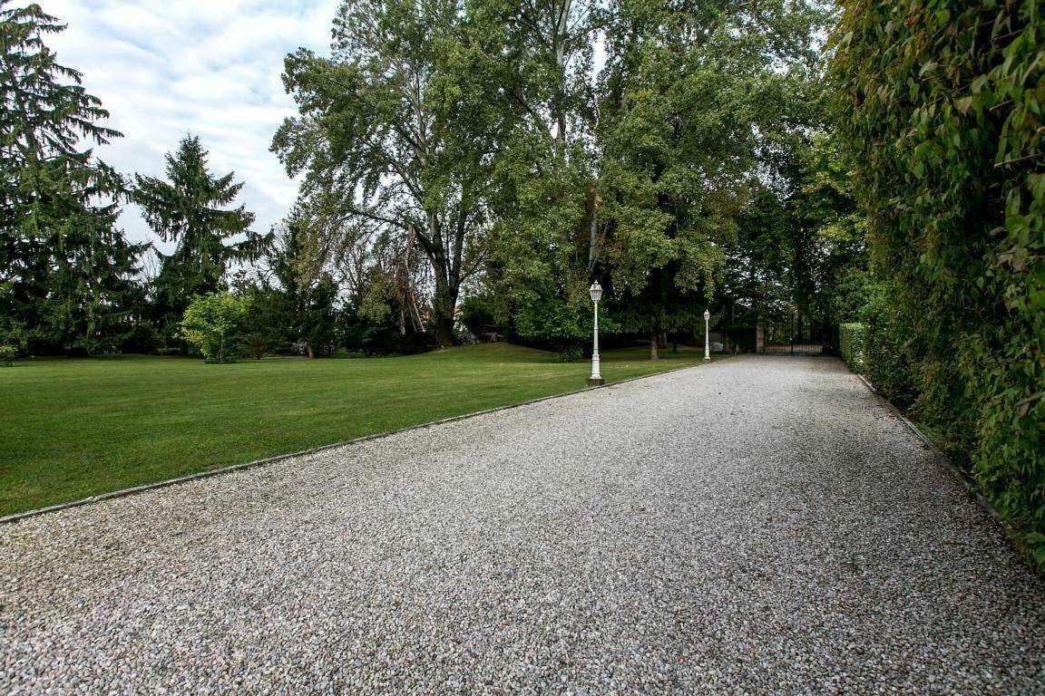 Lange, gepflegte Kiesauffahrt, gesäumt von Laternen und einer dichten Hecke, die zu einem weitläufigen Rasenbereich führt.