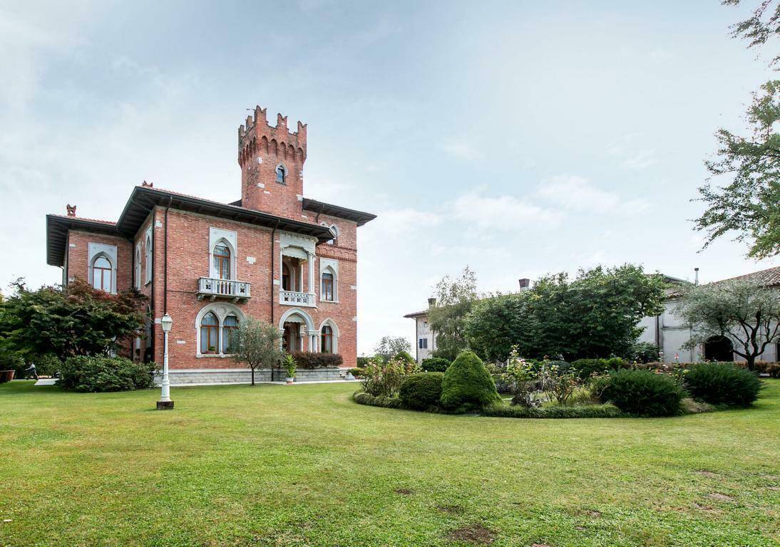 Weitläufiger Rasenbereich vor der historischen Villa mit Ziegelfassade und Turm, umgeben von altem Baumbestand.
