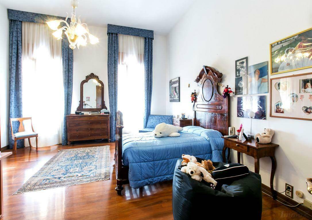 Geräumiges Schlafzimmer mit Holzboden, einem blauen Bett und antiken Holzmöbeln, beleuchtet durch große Fenster.