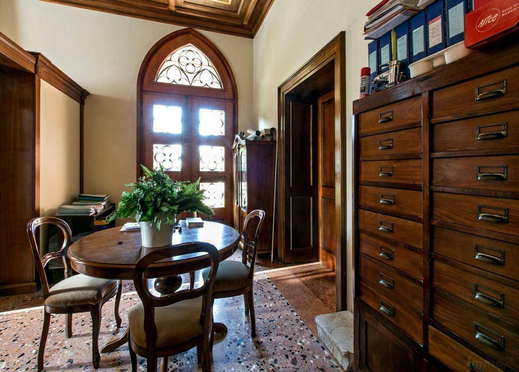 Gemütliches Arbeitszimmer mit Terrazzoboden, einem runden Holztisch und einem großen Holzschrank, beleuchtet durch ein Bogenfenster.