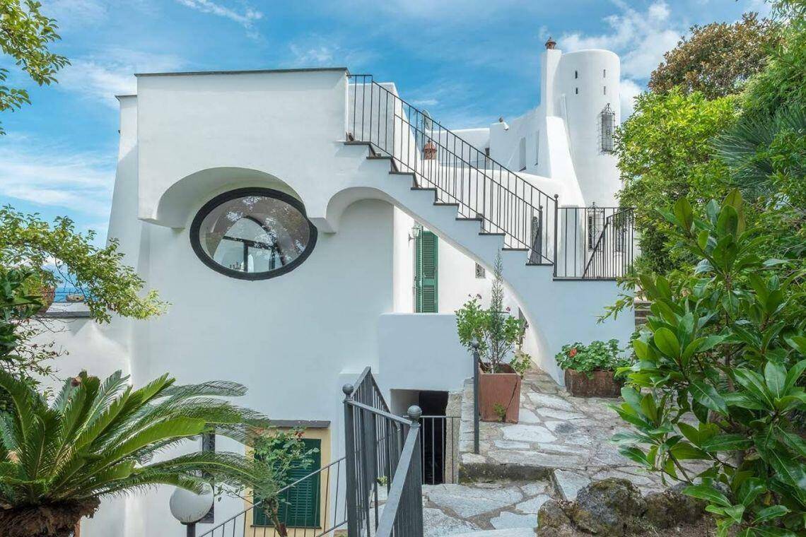 Weiße Fassade mit runden Fenstern, Außentreppe und üppiger mediterraner Bepflanzung.
