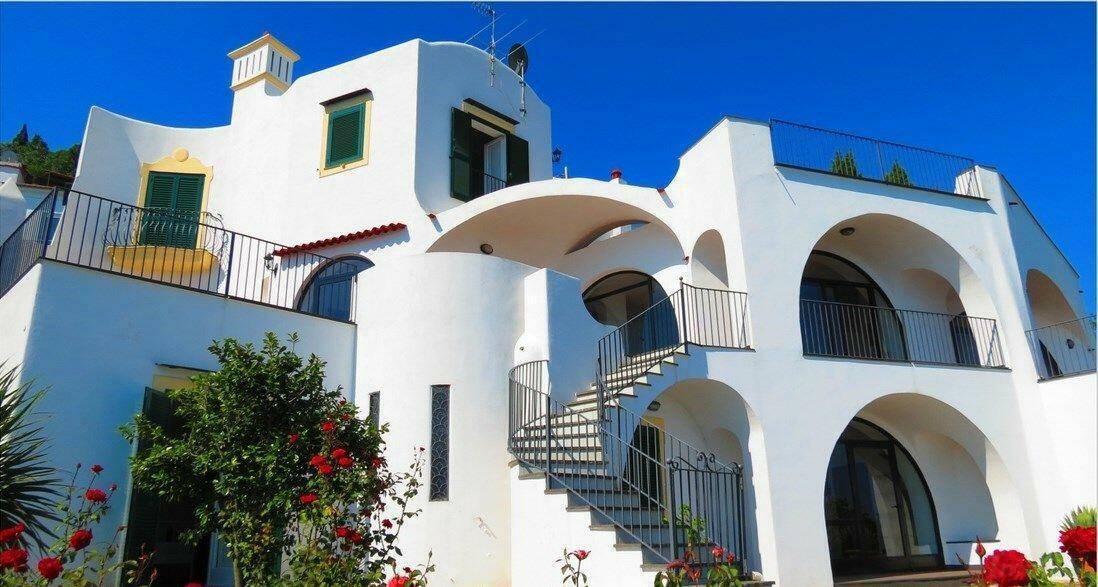 Weiße Villa mit geschwungenen Bögen, Außentreppen und grünen Fensterläden unter blauem Himmel.