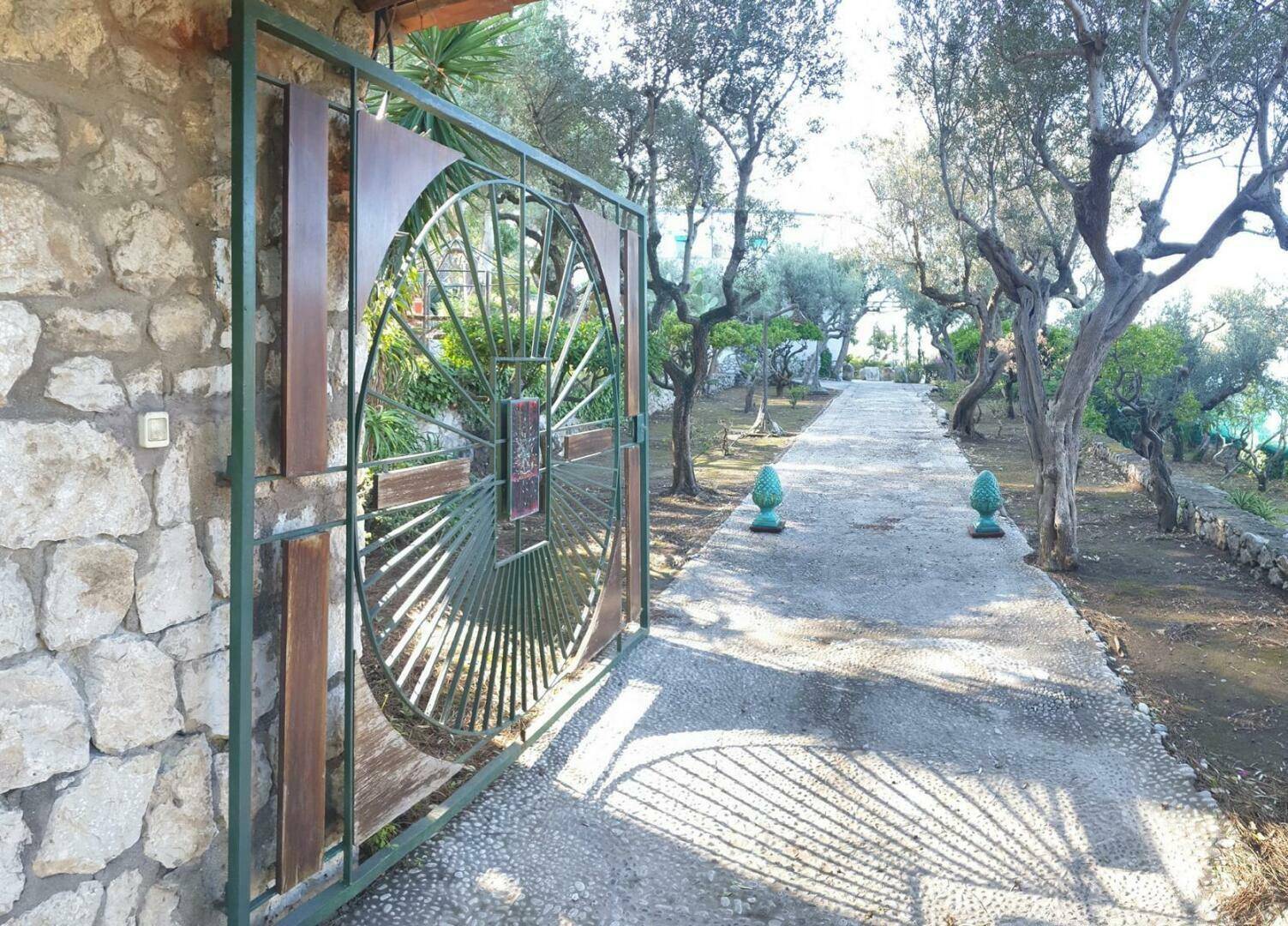 Dekoratives Metalltor in einer Steinmauer, das zu einem von Olivenbäumen gesäumten Gartenweg führt.