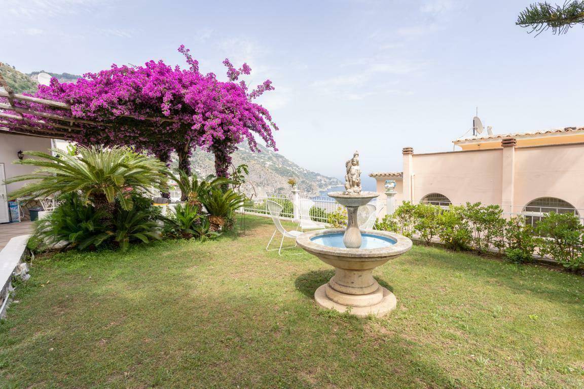 Mediterraner Garten mit Rasenfläche, dekorativem Brunnen und blühender Bougainvillea mit Meerblick.