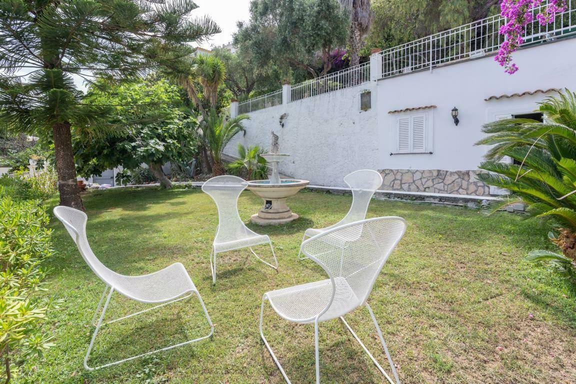 Gepflegter Gartenbereich mit Rasen, dekorativem Brunnen und Sitzgelegenheiten, umgeben von mediterraner Vegetation.